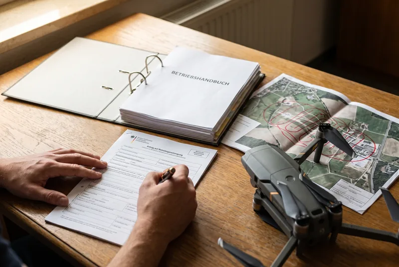 Antragsunterlagen für die Drohnen-Betriebsgenehmigung: Betriebshandbuch, Luftbildkarten mit Fluggebieten und Antragsformular auf einem Schreibtisch